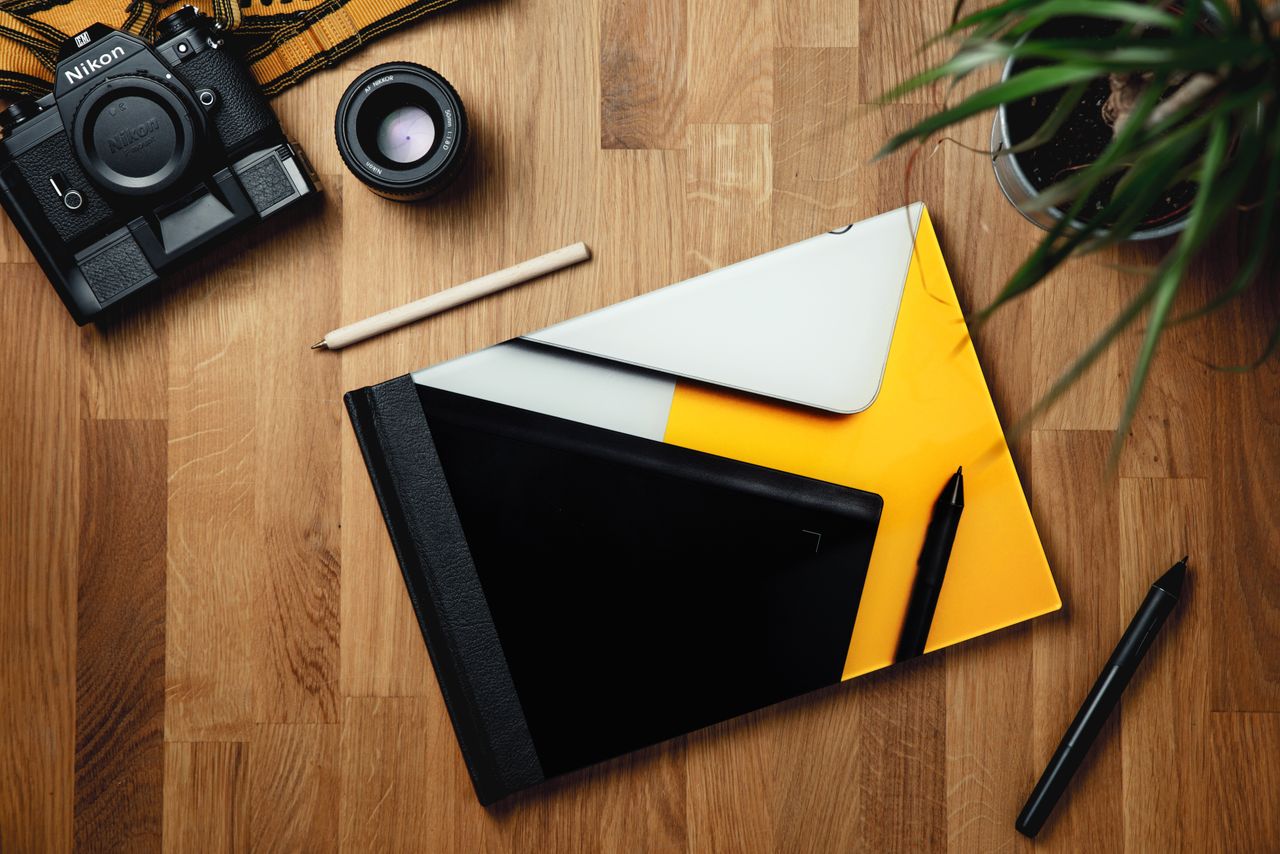 Top view of colorful rectangular notebook surrounded by pens and pot plant composing with professional photo camera and lens on wooden table in bright room